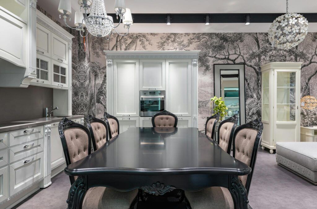 Black table with chairs placed near white cupboards and modern appliances in stylish spacious kitchen with mirror and creative wallpaper